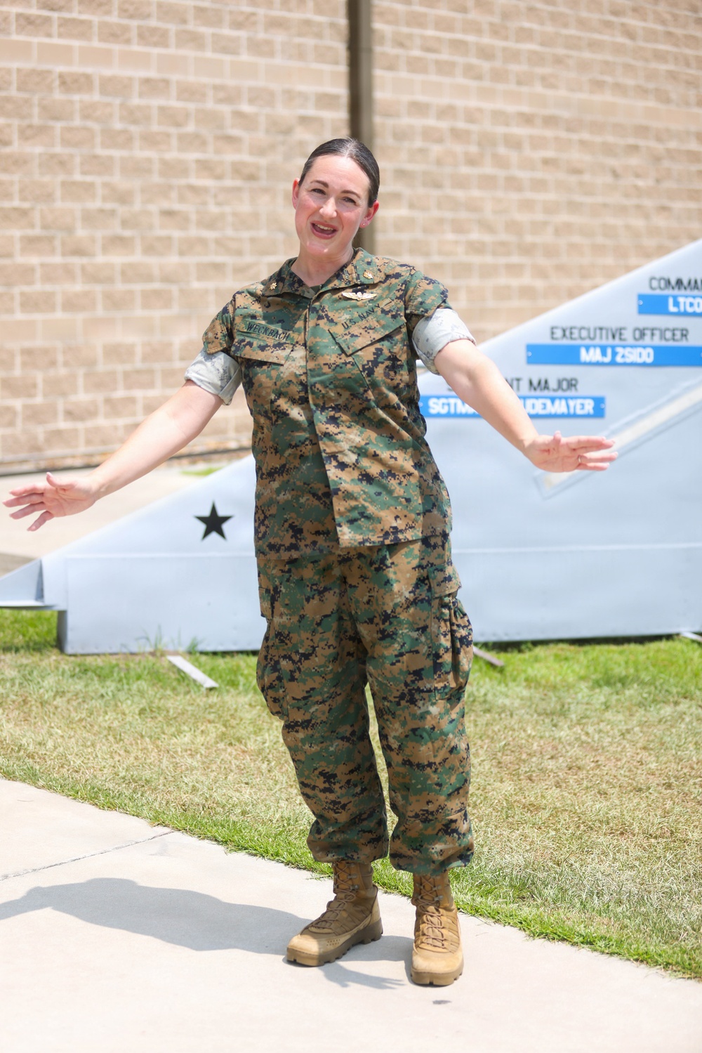 A female military service member wearing Marine Corps MARPAT camouflage utilities stands outdoors on a sidewalk with arms outstretched. She appears to be speaking or presenting in front of a unit command sign board and a brick building. Her name tape reads 'WECKBACH' and she wears U.S. Navy branch tape, indicating a Navy officer attached to a Marine Corps unit.