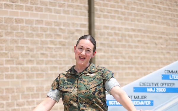 A female military service member wearing Marine Corps MARPAT camouflage utilities stands outdoors on a sidewalk with arms outstretched. She appears to be speaking or presenting in front of a unit command sign board and a brick building. Her name tape reads 'WECKBACH' and she wears U.S. Navy branch tape, indicating a Navy officer attached to a Marine Corps unit.
