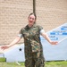 A female military service member wearing Marine Corps MARPAT camouflage utilities stands outdoors on a sidewalk with arms outstretched. She appears to be speaking or presenting in front of a unit command sign board and a brick building. Her name tape reads 'WECKBACH' and she wears U.S. Navy branch tape, indicating a Navy officer attached to a Marine Corps unit.