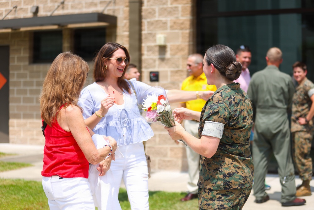 A female service member in U.S. Marine Corps digital camouflage uniform is presenting a bouquet of flowers to two civilian women outside a brick building. The scene appears to be a military ceremony or welcome event, with additional military personnel and civilians visible in the background. The atmosphere is cheerful and celebratory.