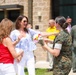A female service member in U.S. Marine Corps digital camouflage uniform is presenting a bouquet of flowers to two civilian women outside a brick building. The scene appears to be a military ceremony or welcome event, with additional military personnel and civilians visible in the background. The atmosphere is cheerful and celebratory.