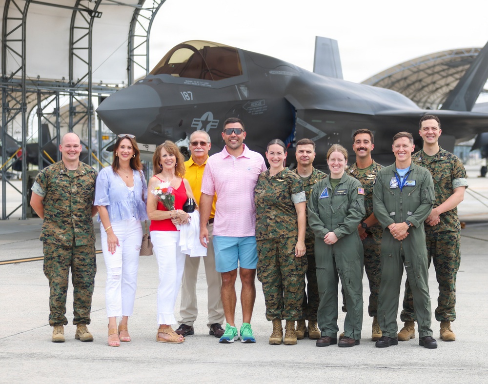 A group of approximately twelve people pose for a photo in front of an F-35 fighter jet on a military airfield tarmac. The group includes both military personnel in Marine Corps camouflage uniforms and flight suits, as well as civilians in casual attire. The setting appears to be a ceremonial or commemorative occasion, possibly a graduation, promotion, or milestone flight event.
