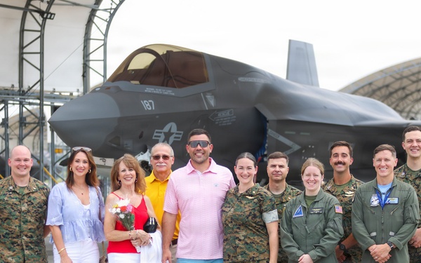 A group of approximately twelve people pose for a photo in front of an F-35 fighter jet on a military airfield tarmac. The group includes both military personnel in Marine Corps camouflage uniforms and flight suits, as well as civilians in casual attire. The setting appears to be a ceremonial or commemorative occasion, possibly a graduation, promotion, or milestone flight event.