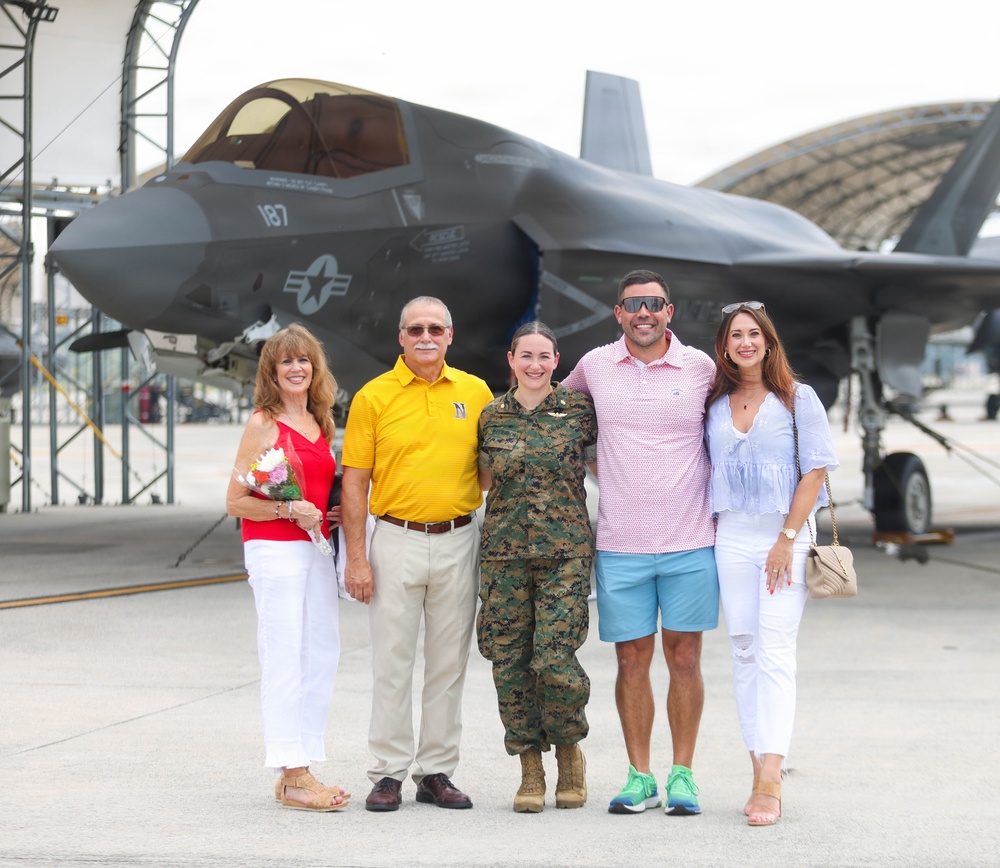 A group of five people pose for a photo on a military airfield tarmac in front of an F-35 stealth fighter jet marked with the number 187 and a U.S. military star insignia. One person in the center is wearing U.S. Marine Corps woodland camouflage uniform, while the other four are dressed in civilian clothing. The setting appears to be a military base with aircraft hangars visible in the background, suggesting a celebratory occasion such as a graduation, promotion, or homecoming.