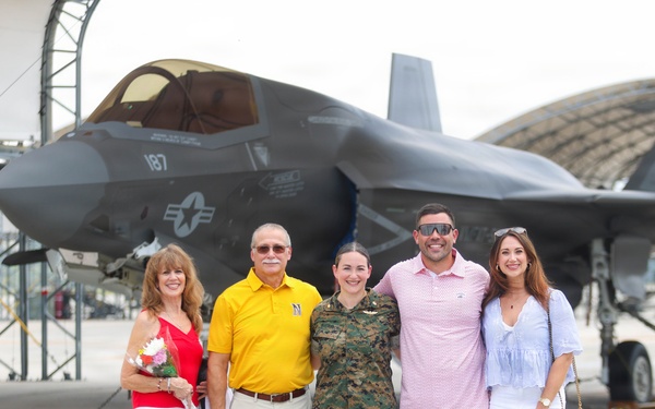 A group of five people pose for a photo on a military airfield tarmac in front of an F-35 stealth fighter jet marked with the number 187 and a U.S. military star insignia. One person in the center is wearing U.S. Marine Corps woodland camouflage uniform, while the other four are dressed in civilian clothing. The setting appears to be a military base with aircraft hangars visible in the background, suggesting a celebratory occasion such as a graduation, promotion, or homecoming.