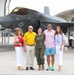 A group of five people pose for a photo on a military airfield tarmac in front of an F-35 stealth fighter jet marked with the number 187 and a U.S. military star insignia. One person in the center is wearing U.S. Marine Corps woodland camouflage uniform, while the other four are dressed in civilian clothing. The setting appears to be a military base with aircraft hangars visible in the background, suggesting a celebratory occasion such as a graduation, promotion, or homecoming.