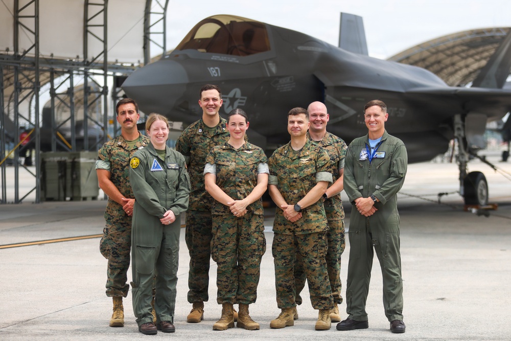 Seven military personnel pose for a group photo on a flight line in front of an F-35 stealth fighter jet marked with the number '187' and a U.S. Air Force star insignia. The group consists of individuals wearing U.S. Marine Corps MARPAT camouflage uniforms and olive green flight suits. The setting appears to be a military air base with hangars visible in the background.