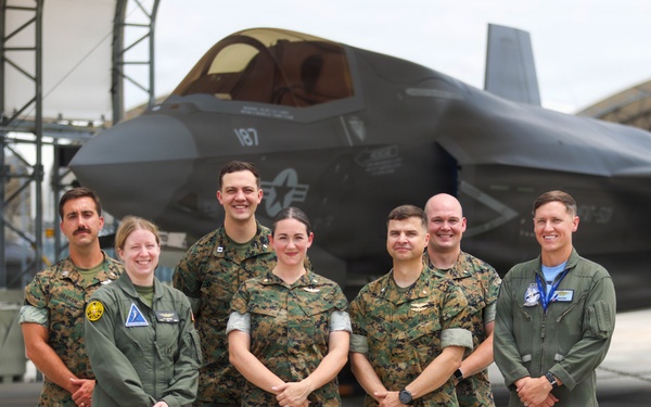 Seven military personnel pose for a group photo on a flight line in front of an F-35 stealth fighter jet marked with the number '187' and a U.S. Air Force star insignia. The group consists of individuals wearing U.S. Marine Corps MARPAT camouflage uniforms and olive green flight suits. The setting appears to be a military air base with hangars visible in the background.