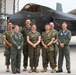 Seven military personnel pose for a group photo on a flight line in front of an F-35 stealth fighter jet marked with the number '187' and a U.S. Air Force star insignia. The group consists of individuals wearing U.S. Marine Corps MARPAT camouflage uniforms and olive green flight suits. The setting appears to be a military air base with hangars visible in the background.