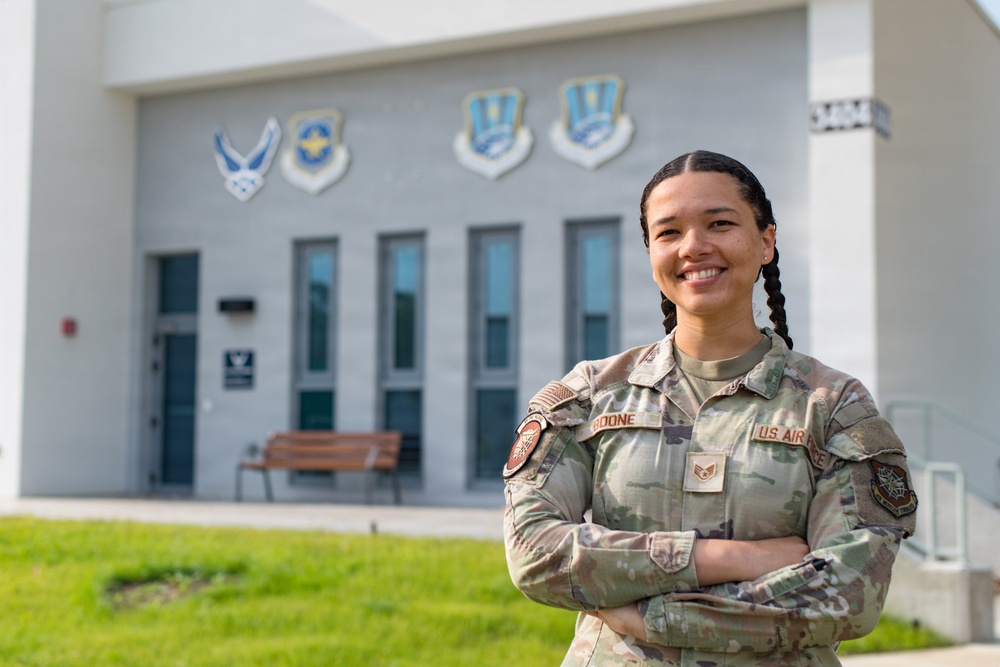 A U.S. Air Force service member named Boone, wearing Operational Camouflage Pattern (OCP) uniform, stands with arms crossed in front of a military building. The individual holds the rank of Technical Sergeant, as indicated by the chevron patch visible on the uniform. The building behind displays multiple U.S. Air Force unit emblems/shields on its facade.