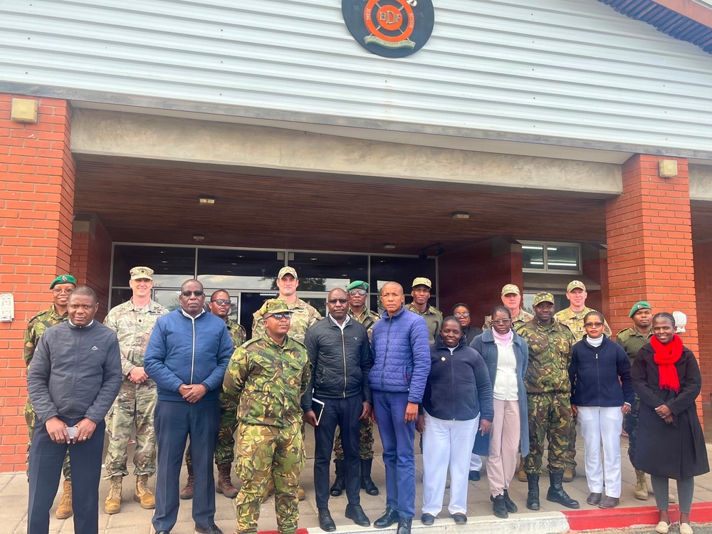 A group of approximately 20 individuals poses for a formal group photograph in front of a building bearing a circular emblem with the letters 'BDF' (Botswana Defence Force). The group consists of a mix of military personnel in camouflage uniforms and civilians in casual attire. The gathering appears to represent a joint or bilateral military meeting or visit.