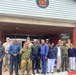 A group of approximately 20 individuals poses for a formal group photograph in front of a building bearing a circular emblem with the letters 'BDF' (Botswana Defence Force). The group consists of a mix of military personnel in camouflage uniforms and civilians in casual attire. The gathering appears to represent a joint or bilateral military meeting or visit.