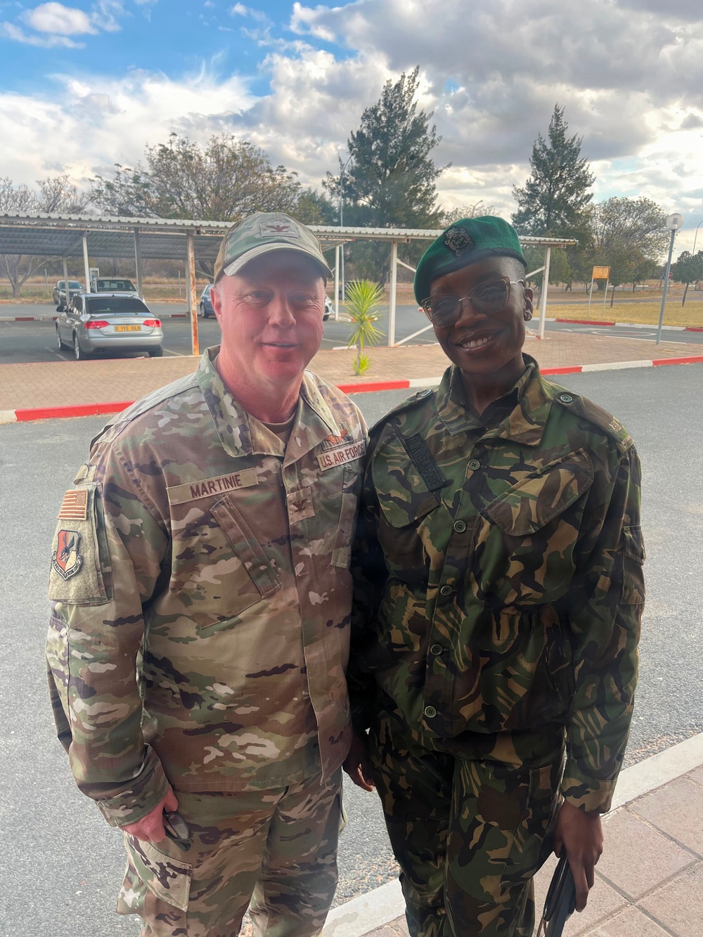 Two military personnel in different uniforms pose together outdoors at what appears to be a military installation or base. The individual on the left wears a U.S. Air Force OCP (Operational Camouflage Pattern) uniform with the name tape 'MARTINIE' and a U.S. flag patch, while the individual on the right wears a DPM (Disruptive Pattern Material) camouflage uniform with a green beret. The setting appears to be a paved area with a parking lot and trees visible in the background.