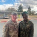 Two military personnel in different uniforms pose together outdoors at what appears to be a military installation or base. The individual on the left wears a U.S. Air Force OCP (Operational Camouflage Pattern) uniform with the name tape 'MARTINIE' and a U.S. flag patch, while the individual on the right wears a DPM (Disruptive Pattern Material) camouflage uniform with a green beret. The setting appears to be a paved area with a parking lot and trees visible in the background.