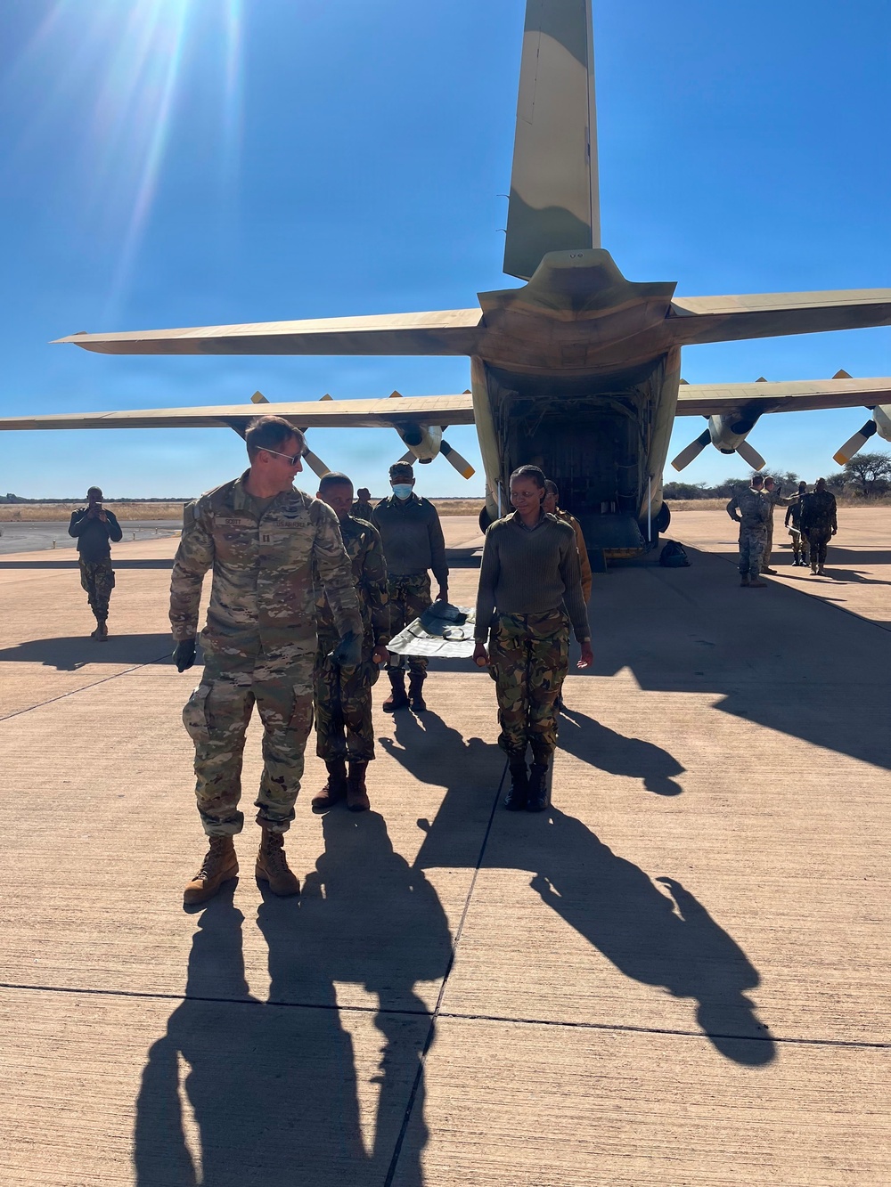 Military personnel from what appears to be two different armed forces are gathered on an airfield tarmac near a large military transport aircraft with its rear cargo ramp open. One individual in US Army MultiCam/OCP uniform is visible alongside personnel wearing a different camouflage pattern, suggesting a joint or cooperative military exercise or operation. The setting appears to be an arid or semi-arid region based on the dry vegetation visible in the background.