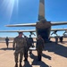 Military personnel from what appears to be two different armed forces are gathered on an airfield tarmac near a large military transport aircraft with its rear cargo ramp open. One individual in US Army MultiCam/OCP uniform is visible alongside personnel wearing a different camouflage pattern, suggesting a joint or cooperative military exercise or operation. The setting appears to be an arid or semi-arid region based on the dry vegetation visible in the background.