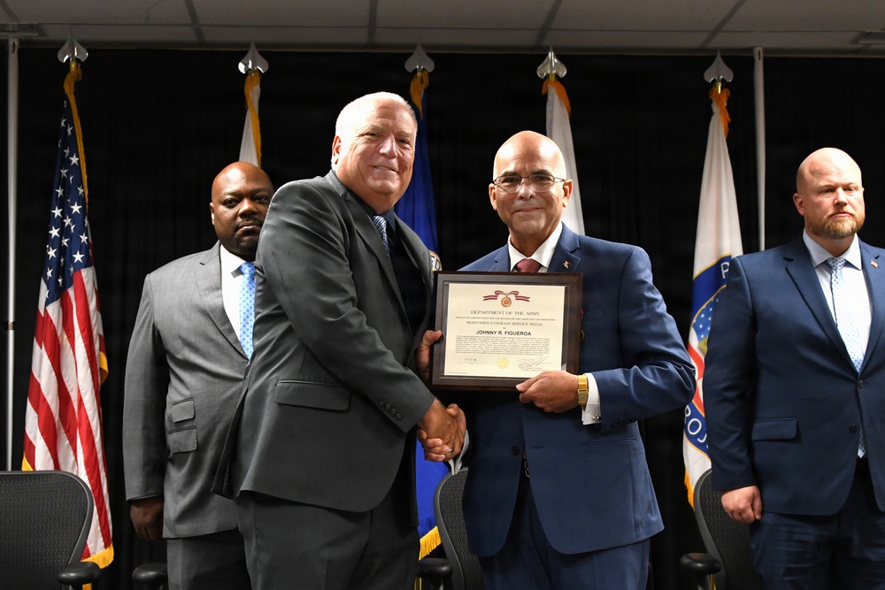 A formal award ceremony is taking place in what appears to be a government or military facility. Two men in suits are shaking hands while jointly holding a framed Department of the Army certificate awarded to 'Johnny R. Figueroa.' Two additional men stand in the background flanking the central figures.