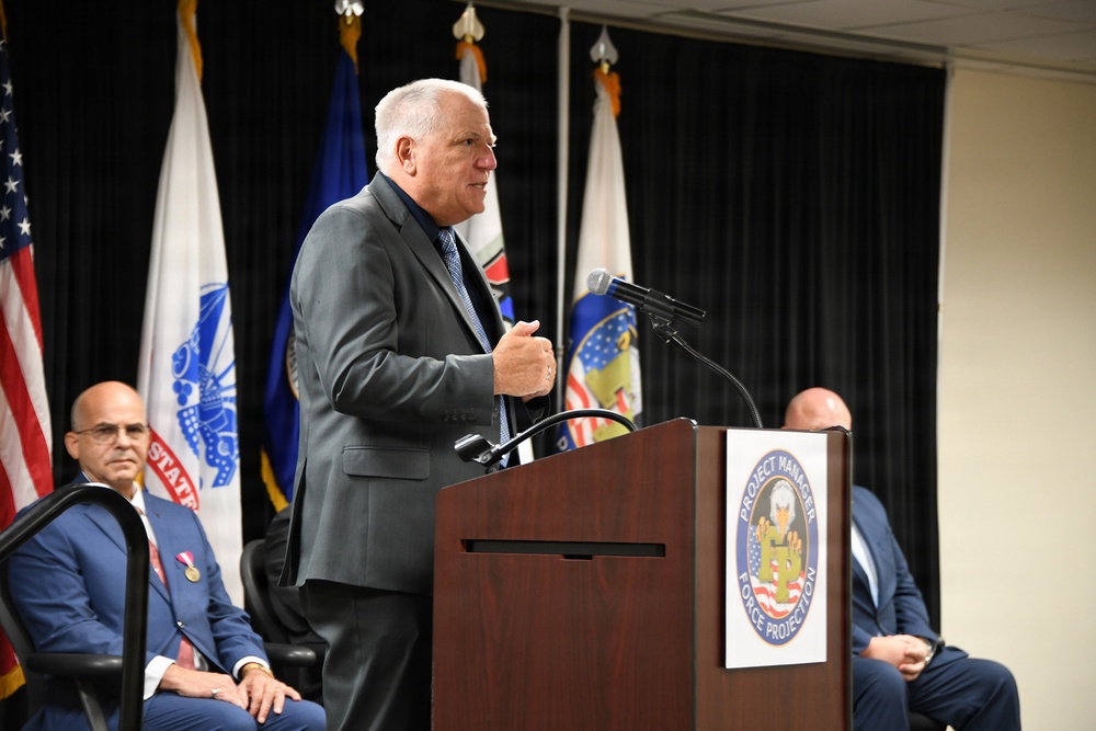 A man in a gray suit is delivering a speech at a podium bearing the seal of 'Project Manager Force Projection,' a U.S. Army program office. Multiple U.S. military and government flags are displayed in the background, indicating an official ceremony or formal event. Two other men in suits are seated behind the speaker, one visibly wearing a medal.