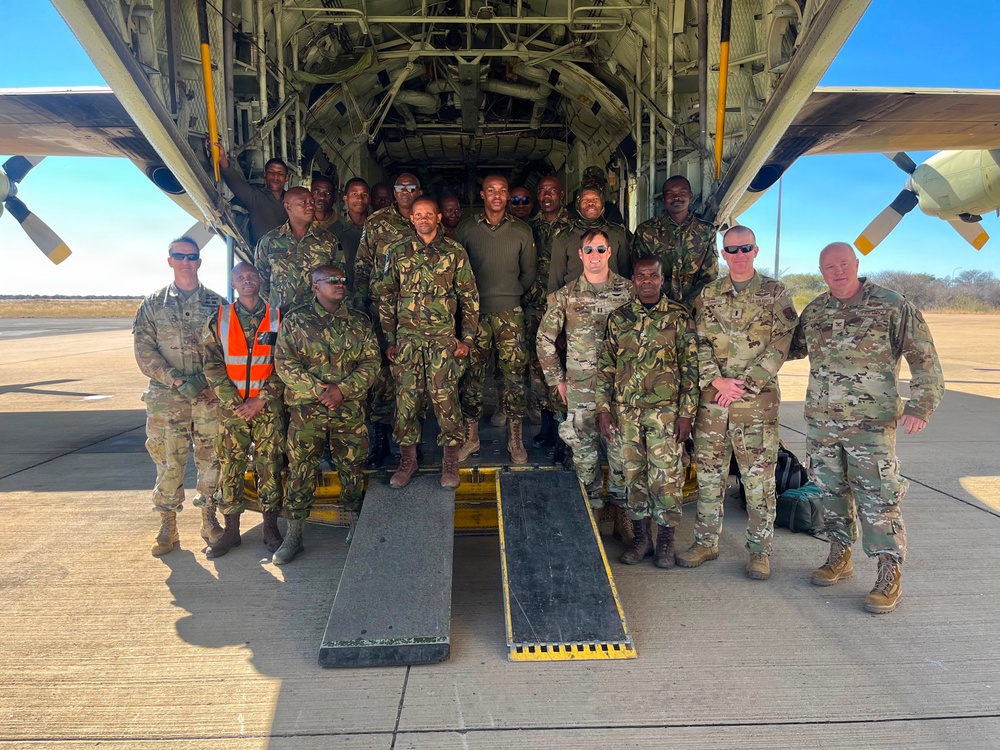 A group of military personnel from at least two different forces pose for a group photo in front of the open rear cargo ramp of a large military transport aircraft, likely a C-130 Hercules. The group includes soldiers wearing US Army MultiCam/OCP uniforms and others wearing a different camouflage pattern consistent with an African military force. The setting appears to be a tarmac at a military airfield in an arid environment.