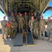 A group of military personnel from at least two different forces pose for a group photo in front of the open rear cargo ramp of a large military transport aircraft, likely a C-130 Hercules. The group includes soldiers wearing US Army MultiCam/OCP uniforms and others wearing a different camouflage pattern consistent with an African military force. The setting appears to be a tarmac at a military airfield in an arid environment.