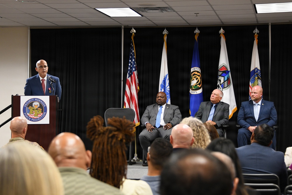 A formal ceremony or official event is taking place at a 'Project Manager Force Projection' podium, with a speaker addressing an audience. Three dignitaries are seated on a stage behind the speaker, flanked by multiple flags including the U.S. flag and what appear to be U.S. Army Corps of Engineers and other organizational flags. An audience of approximately 20+ attendees is visible from behind in the foreground.