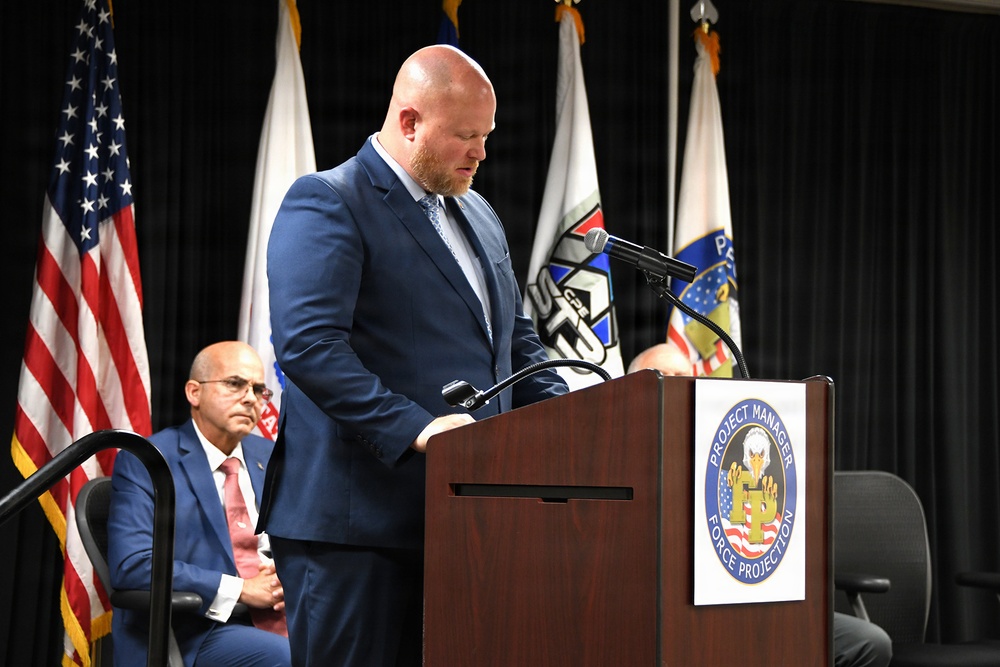 A bald man in a navy blue suit is speaking at a wooden podium bearing the seal of 'Project Manager Force Projection,' a U.S. Army program office. He appears to be looking down, likely reading from notes, at what appears to be an official ceremony or briefing. A second man in a blue suit with a red tie is seated in the background.