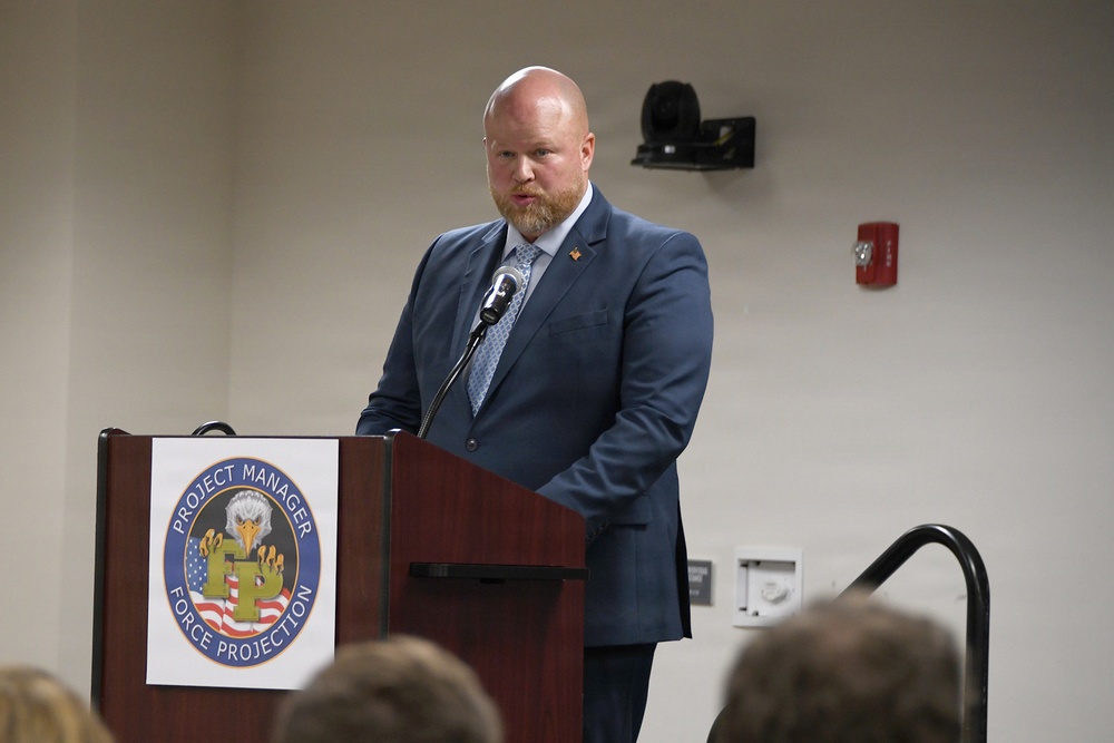 A man in a blue suit is speaking at a wooden podium bearing the seal of 'Project Manager Force Protection,' which features an eagle and an American flag design. The setting appears to be a formal briefing or conference room with an audience visible in the foreground. The speaker has a reddish beard and is wearing a patterned tie with a lapel pin.