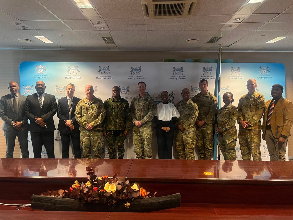 A group of approximately 13 individuals pose for a formal group photograph in front of a Republic of Botswana Ministry of Health branded backdrop. The group includes both military personnel in camouflage uniforms and civilians in business attire. The setting appears to be a conference or meeting room, suggesting an official diplomatic or cooperative health-related engagement.