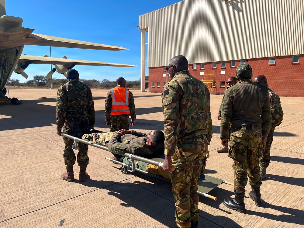 Military personnel in camouflage uniforms are conducting what appears to be a medical evacuation (MEDEVAC) exercise or operation on an airfield tarmac. A person on a stretcher is being carried toward a military transport aircraft, likely a C-130 Hercules, visible on the left side of the frame. A large aircraft hangar and brick building are visible in the background.