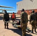 Military personnel in camouflage uniforms are conducting what appears to be a medical evacuation (MEDEVAC) exercise or operation on an airfield tarmac. A person on a stretcher is being carried toward a military transport aircraft, likely a C-130 Hercules, visible on the left side of the frame. A large aircraft hangar and brick building are visible in the background.