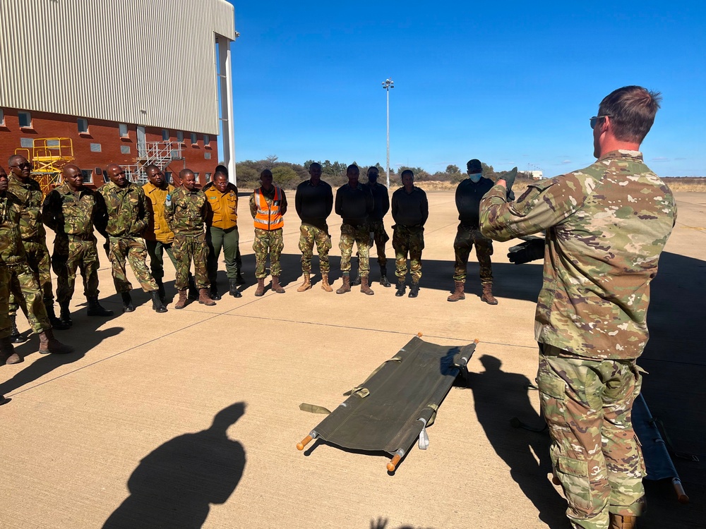 A military instructor in MultiCam/OCP camouflage uniform is conducting a training session on an airfield tarmac, addressing a group of approximately 15 personnel in mixed military and civilian attire. A folded military litter/stretcher is placed on the ground in front of the group, suggesting medical or casualty evacuation training. The setting appears to be an African military installation based on the architecture and vegetation visible in the background.