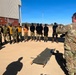 A military instructor in MultiCam/OCP camouflage uniform is conducting a training session on an airfield tarmac, addressing a group of approximately 15 personnel in mixed military and civilian attire. A folded military litter/stretcher is placed on the ground in front of the group, suggesting medical or casualty evacuation training. The setting appears to be an African military installation based on the architecture and vegetation visible in the background.