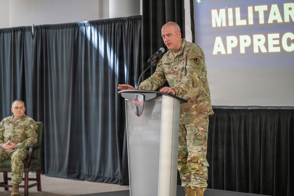 A U.S. Army officer in OCP (Operational Camouflage Pattern) uniform is speaking at a podium during what appears to be a Military Appreciation event. A second uniformed soldier is seated in the background to the left. The setting appears to be an indoor venue with black curtain backdrops and a projected presentation screen.