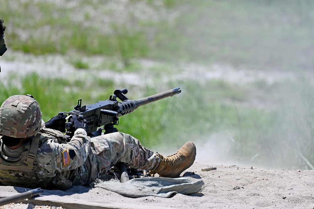 A U.S. Army soldier in OCP (Operational Camouflage Pattern) uniform is firing a heavy machine gun from a prone position on a sandy range. The weapon appears to be an M2 .50 caliber machine gun mounted on a bipod or tripod. A spent brass casing is visible ejecting near the soldier's feet, and muzzle blast dust is visible downrange.