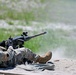 A U.S. Army soldier in OCP (Operational Camouflage Pattern) uniform is firing a heavy machine gun from a prone position on a sandy range. The weapon appears to be an M2 .50 caliber machine gun mounted on a bipod or tripod. A spent brass casing is visible ejecting near the soldier's feet, and muzzle blast dust is visible downrange.