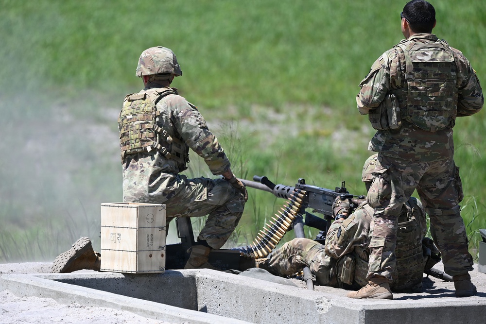 Three soldiers in MultiCam/OCP camouflage uniforms are positioned at a concrete barrier during what appears to be a live-fire training exercise. One soldier is in a prone position operating a belt-fed machine gun, while two others provide support and observation. A wooden ammunition crate is visible nearby, and a belt of linked ammunition feeds into the weapon.