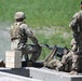 Three soldiers in MultiCam/OCP camouflage uniforms are positioned at a concrete barrier during what appears to be a live-fire training exercise. One soldier is in a prone position operating a belt-fed machine gun, while two others provide support and observation. A wooden ammunition crate is visible nearby, and a belt of linked ammunition feeds into the weapon.