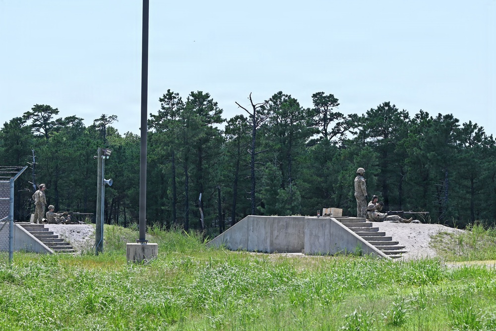 The image shows military personnel in camouflage uniforms positioned at what appears to be a firing range or tactical training facility with concrete berms and stepped embankments. Several soldiers are visible in various positions — standing, kneeling, and prone — suggesting an active training exercise. The facility is surrounded by pine trees consistent with a southeastern U.S. military installation.