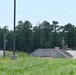 The image shows military personnel in camouflage uniforms positioned at what appears to be a firing range or tactical training facility with concrete berms and stepped embankments. Several soldiers are visible in various positions — standing, kneeling, and prone — suggesting an active training exercise. The facility is surrounded by pine trees consistent with a southeastern U.S. military installation.