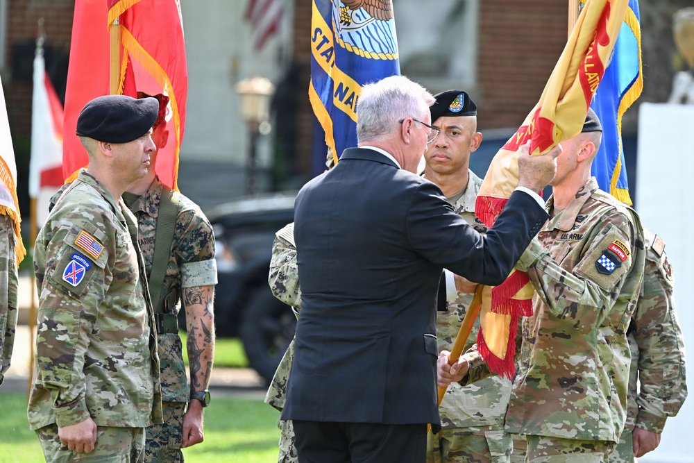 A military ceremony is taking place outdoors, likely a change of command or unit activation/inactivation ceremony. A civilian in a dark suit is passing or presenting a unit guidon (flag) to a U.S. Army soldier in multicam uniform. Personnel from multiple military branches, including Army and Marine Corps, are present as participants.