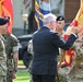 A military ceremony is taking place outdoors, likely a change of command or unit activation/inactivation ceremony. A civilian in a dark suit is passing or presenting a unit guidon (flag) to a U.S. Army soldier in multicam uniform. Personnel from multiple military branches, including Army and Marine Corps, are present as participants.