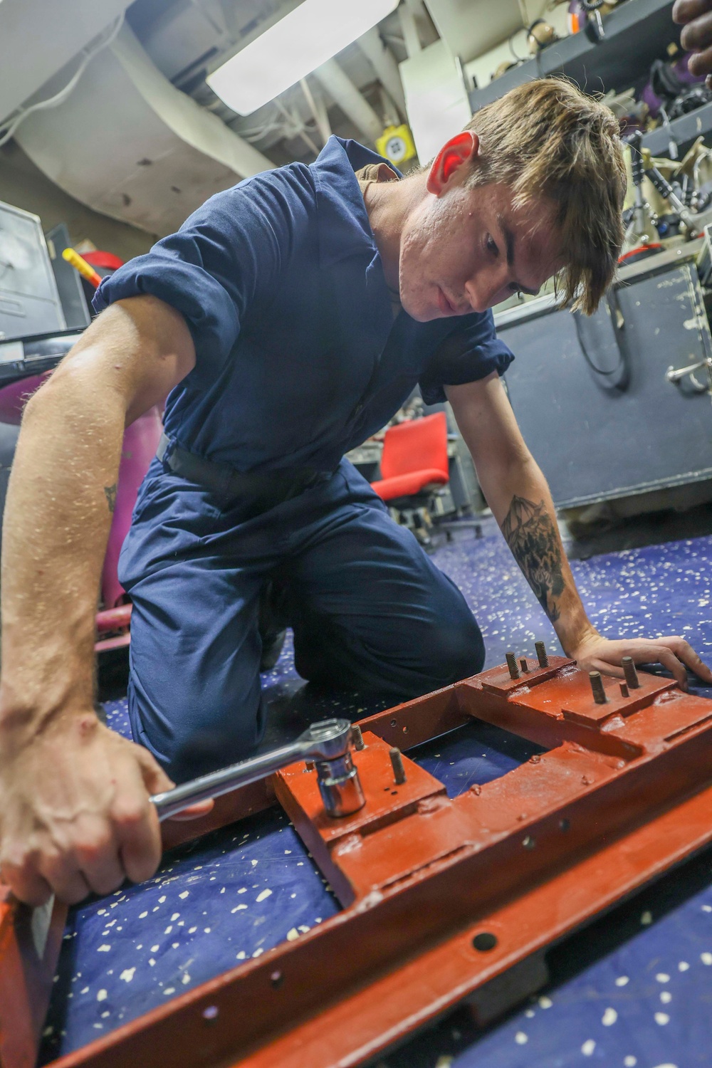 A person wearing a navy blue coverall uniform is kneeling on a blue speckled deck floor, using a ratchet wrench to work on a red-painted metal component. The setting appears to be an interior workspace aboard a military vessel, likely a U.S. Navy ship based on the coverall style and environment. The individual has a visible tattoo on their right forearm and is focused on the mechanical task.