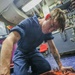 A person wearing a navy blue coverall uniform is kneeling on a blue speckled deck floor, using a ratchet wrench to work on a red-painted metal component. The setting appears to be an interior workspace aboard a military vessel, likely a U.S. Navy ship based on the coverall style and environment. The individual has a visible tattoo on their right forearm and is focused on the mechanical task.