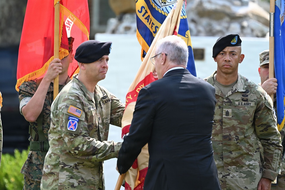 A military ceremony is taking place, likely a change of command or activation ceremony, where a guidon or unit colors are being passed between a U.S. Army officer and a civilian official in a dark suit. Multiple service members in various uniforms are present as color guards, holding flags including what appears to be a U.S. Army unit flag and other organizational colors. The soldier receiving or passing the colors wears a 10th Mountain Division patch on his left shoulder.
