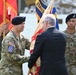 A military ceremony is taking place, likely a change of command or activation ceremony, where a guidon or unit colors are being passed between a U.S. Army officer and a civilian official in a dark suit. Multiple service members in various uniforms are present as color guards, holding flags including what appears to be a U.S. Army unit flag and other organizational colors. The soldier receiving or passing the colors wears a 10th Mountain Division patch on his left shoulder.