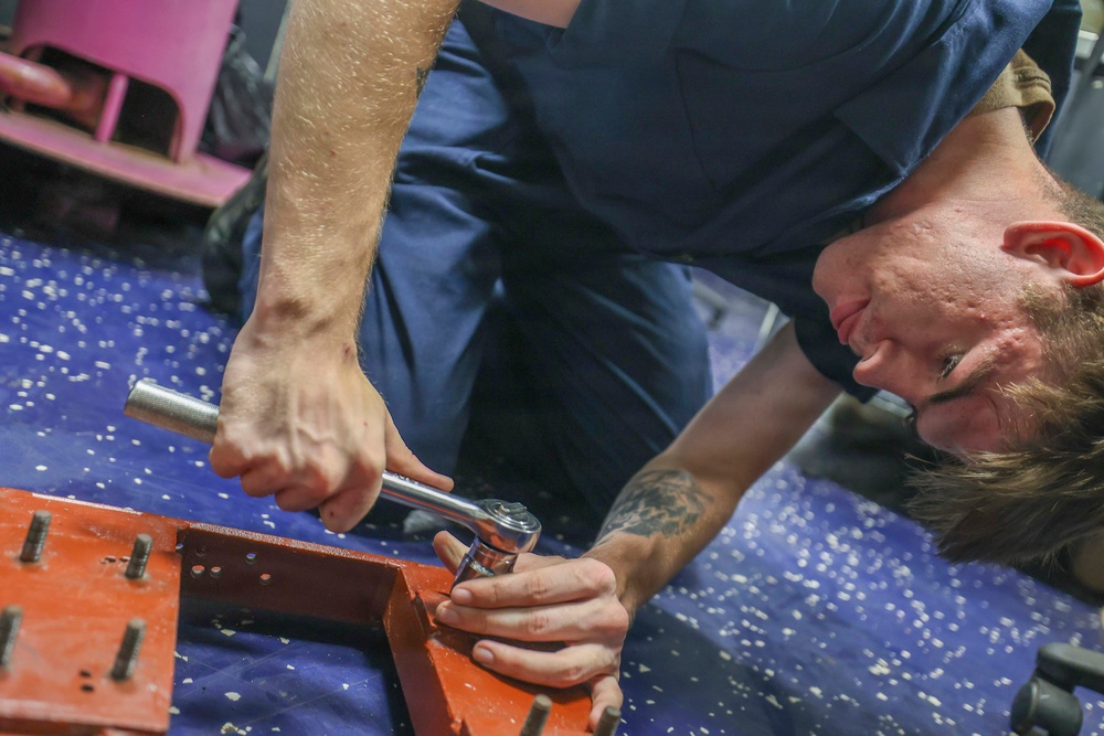 A young man wearing a navy blue work uniform is lying on a blue mat, using a ratchet wrench to work on a red metal frame or bracket. He is focused intently on the task, positioning his face close to the work area for precision. A tattoo is visible on his forearm, and pink equipment is partially visible in the background.