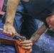 A young man wearing a navy blue work uniform is lying on a blue mat, using a ratchet wrench to work on a red metal frame or bracket. He is focused intently on the task, positioning his face close to the work area for precision. A tattoo is visible on his forearm, and pink equipment is partially visible in the background.