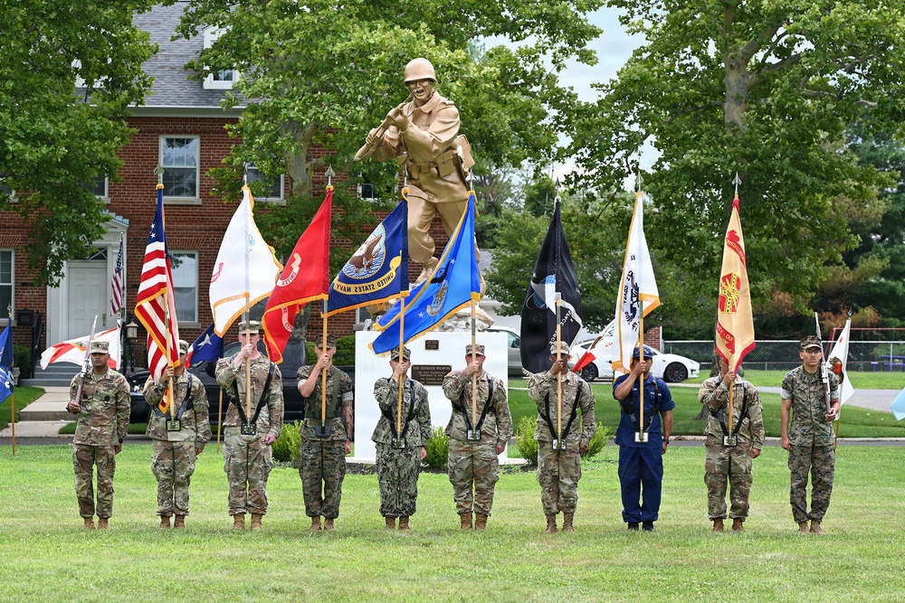 A military color guard ceremony is taking place outdoors on a grassy area, with service members from multiple branches holding various flags and guidons. Behind them stands a large golden/bronze statue of a soldier in combat pose on a white pedestal. The setting appears to be a military installation with brick buildings and mature trees in the background.