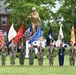 A military color guard ceremony is taking place outdoors on a grassy area, with service members from multiple branches holding various flags and guidons. Behind them stands a large golden/bronze statue of a soldier in combat pose on a white pedestal. The setting appears to be a military installation with brick buildings and mature trees in the background.