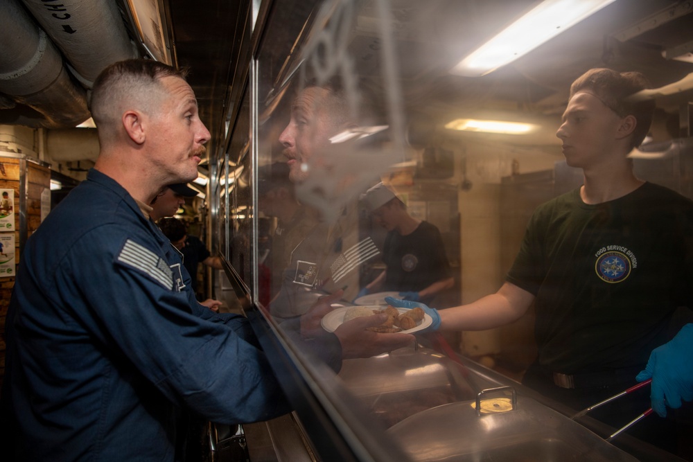 The image shows a military food service line, likely aboard a naval vessel, where personnel in a 'Food Service Division' shirt are serving food to uniformed service members. A person in a blue flight suit with a U.S. flag patch is receiving a plate of food through a sneeze guard. The setting appears to be a ship's galley or mess hall.